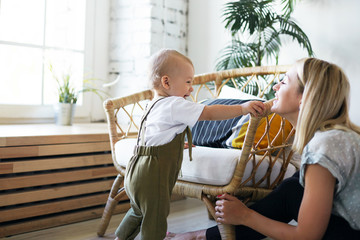 Parenthood, maternity and childcare concept. Indoor image of casually dressed young female babysitter playing with happy toddler who is standing upright, holding on to armchair, smiling excitedly