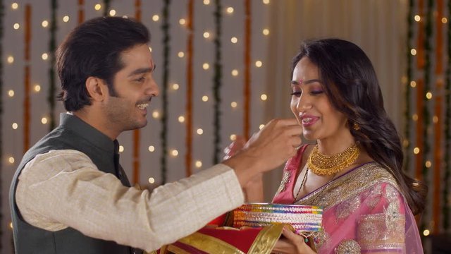 Young attractive couple preforming Karwa Chauth traditions - Indian festival. Handsome husband offering sweets to her pretty wife to complete her fast successfully on the occasion of Karwa Chauth
