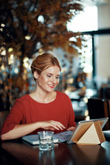 Smiling young woman sitting at restaurant table and making video call when waiting for order