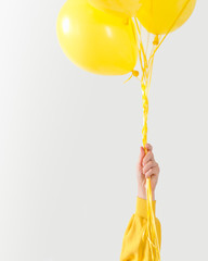 A hand in a yellow hoodie holds balloons of the same color by a bunch of strings and rises upwards, a concept of joyful state, friendship and support. Light background.
