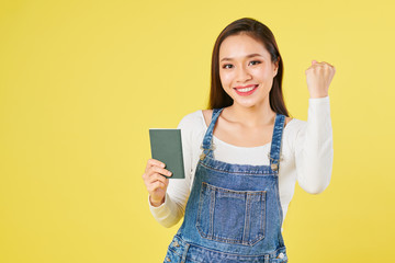 Portrait of happy young Asian woman in denim overall making fist bump and showing passport with visa in it, isolated on yellow