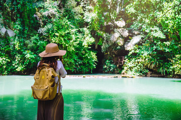 Girl with a yellow backpack is walking travel nature. The young woman is standing, watching the beautiful emerald brook at the shoulder down from the waterfall in the deep forest.