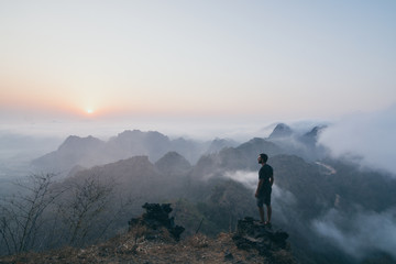 Man standing on the rock overlooking tropical mountains at sunrise foggy morning in Hpa-an, Myanmar