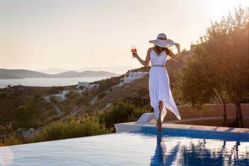 Elegante Frau in weißem Sommerkleid steht am Pool und genießt den Sonnenuntergang mit einem...