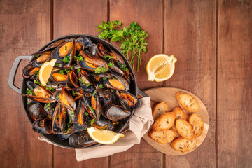 Marinara mussels, moules mariniere, with toasted bread, parsley, and lemon slices, in a cooking pot, overhead shot on a dark rustic wooden background