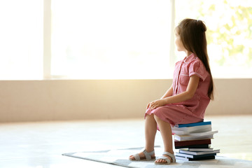 Cute little girl sitting on stack of books at home