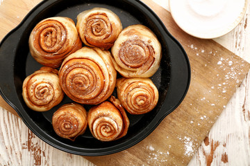 Fototapeta premium Freshly baked cinnamon rolls in black baking pan with powdered sugar on wooden board, overhead view