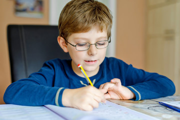 Portrait of cute school kid boy wearing glasses at home making homework. Little concentrated child writing with colorful pencils, indoors. Elementary school and education
