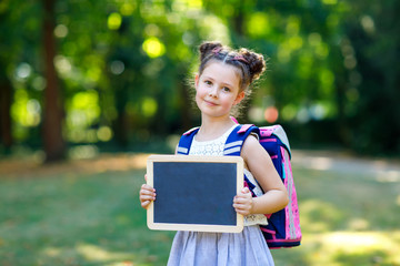 Happy little kid girl standing with desk and backpack or satchel. Schoolkid on first day of elementary class. Healthy adorable child outdoors, in green park. Copyspace empty desk