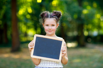 Happy little kid girl holding empty chalk desk in hands. Schoolkid on first day of elementary class. Healthy adorable child outdoors, in green park. dream profession