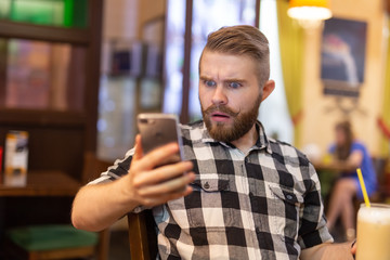 Shocked young hipster man drinking cocoa from a straw and looking at messages in social networks while sitting at a table in a cafe. The concept of chatting and social networks.