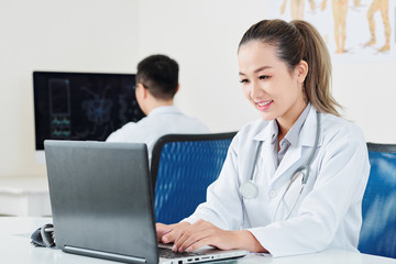 Fototapeta na wymiar Portrait of smiling pretty Vietnamese doctor entering patient information in computer program