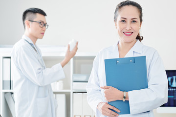 Cheerful mature Vietnamese doctor with clipboard smiling and looking at camera