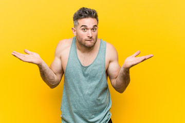 Young fitness man against a yellow background confused and doubtful shrugging shoulders to hold a...