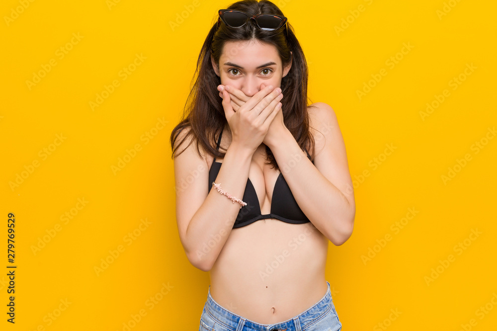 Wall mural young brunette woman wearing a bikini against yellow background shocked covering mouth with hands.