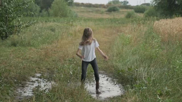 Beautiful smiling girl jumping in very muddy puddle on country road, summer nature background after rain