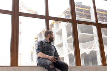 Obraz na płótnie Canvas Young handsome man hipster sitting on the windowsill near a large window with a smartphone in his hands and looking at the construction site at home. Novice architect concept.