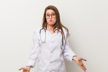 Young doctor woman against a white wall shrugs shoulders and open eyes confused.