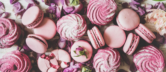 Flat-lay of sweet pink macaron cookies, lilac marshmallows and spring flowers and petals over wooden background, top view, wide composition, close-up. Food texture, background and wallpaper