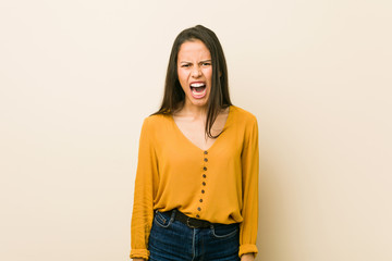 Young hispanic woman against a beige background screaming very angry and aggressive.