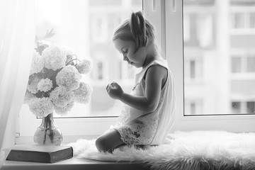 A little girl is sitting on the windowsill. A bouquet of flowers in a vase by the window and a girl...