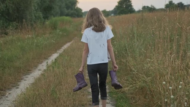 Little girl walking on a rural road with boots in her hands. Happy child in dirty clothes, blurred playing in puddle, back view, summer nature background