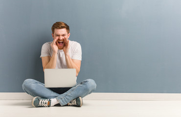 Young redhead student man sitting on the floor shouting something happy to the front. He is holding...