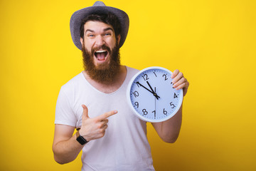 Photo of excited bearded guy with hat, pointing at  white clock over yellow background