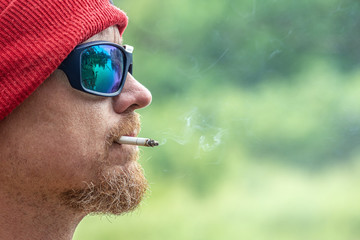 brutal man in black sunglass smoking a cigarette in green forest, outdoor hipster portrait