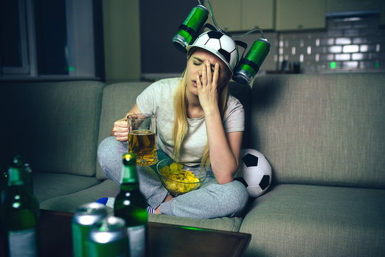 Young Woman Watch Football Game On Tv At Night. Dissapointed Emotional Model Hold Hand On Face. Beer Mug In Hand. Ball Behind Woman On Sofa. Wear Special Hat With Cans On Each Side.