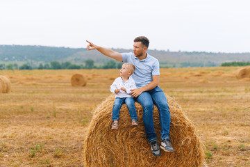 Naklejka na ściany i meble Photo of a dad and his little boy sitting on a round ball of wheat, dad pointing up