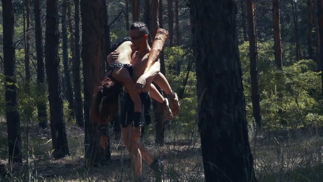 Barbarian man with a bat in hand is carrying an unconscious woman in the forest