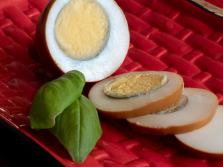 Brown marinated, pickled eggs, sliced egg slices, basil leaves on a red plate, on a light table top