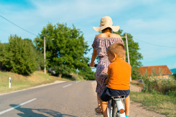 Young woman in a sunny summer day wearing dress riding a bike bicycle on the open road on the asphalt with her small boy kid child son in the back sitting