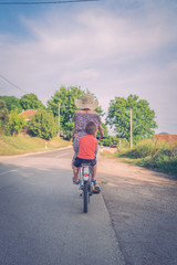 Young woman in a sunny summer day wearing dress riding a bike bicycle on the open road on the asphalt with her small boy kid child son in the back sitting