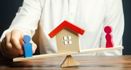 Wooden figures of a man and a woman on scales and a house between them. Division of property by legal means. Clarification of ownership of the housing. Divorce concept. Lawyer. Court. Spouses