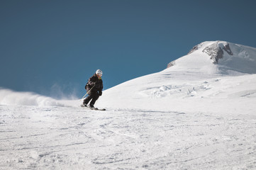 A bearded mature aged male skier in a black ski suit descends along the snowy slope of a ski resort amid two peaks of Mount Elbrus. The concept of sports in adulthood