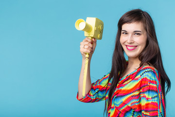 Beautiful positive young brunette woman holding in her hands a model of a yellow camcorder posing on a blue background. Home video recording concept.