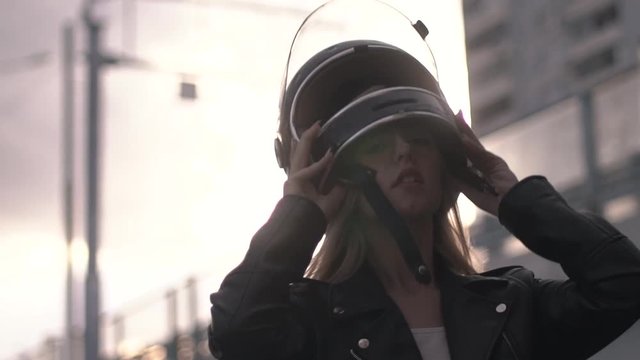 Close-up Of Young Motorcyclist Woman Putting On Retro Helmet In The Outdoors At Sunset. Biker Girl In Leather Jacket Dressing For Trip. Front View, Slow Mo