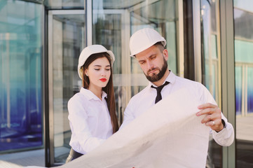 a woman and a man in business clothes and in white construction helmets discuss a construction plan or a contract signature against the background of a modern glass building