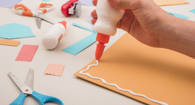 Human hand applying white glue on orange paper with scissor and stapler
