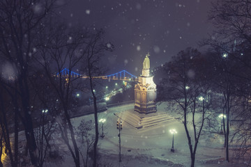 Illuminated bronze statue of Saint Volodymyr in winter Kyiv. Frosted Dnieper river and foot bridge at background.