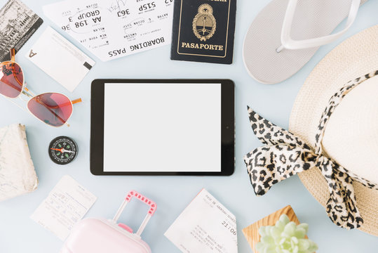 Blank digital tablet surrounded with boarding passes; visiting card; sunglasses; compass; cactus plant; hat; passport; miniature travel bag and flip flops
