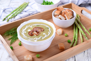 Vegetable cream soup with asparagus and peas garnished with cream and roasted champignons and a bowl of croutons next to it