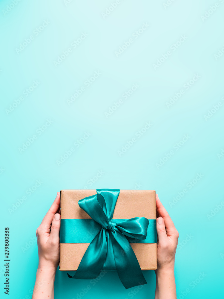 Wall mural Female hands hold big gift box on turquoise blue background, copy space. Caucasian girl hands holding gift box in craft wrapping paper with green satin ribbon. Christmas, New Year or Birthday present