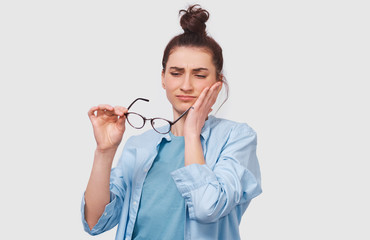 Questioned pretty young woman has thinking expression, looking through her transparent eyeglasses. Doubtful girl wears eyewear can`t make choice, has questioned expression, on white wall