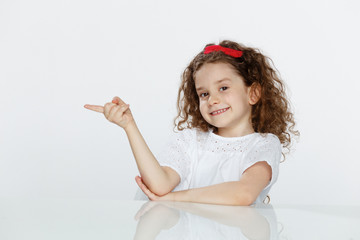 Portrait of a little adorable curly girl, seated at table, showing with finger on direction, over white background.