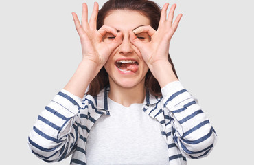 Closeup horizontal studio image of cheerful and funny young woman, showing Ok gestures with both hands, pretending to wear spectacles ot binocular. Pretty female showing tongue.