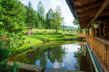 Obraz premium Close up. Small picturesque lake near the wooden house. Surrounded by lush green vegetation and the Carpathian Mountains.