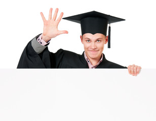 Male graduate student peeking from behind a blank panel, isolated on white background. Handsome graduate guy student in mantle showing blank placard board.
